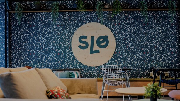 Un área de descanso moderna con un sofá, una mesa redonda y una silla. En la pared, se observa un letrero circular con las letras 'SLO'.