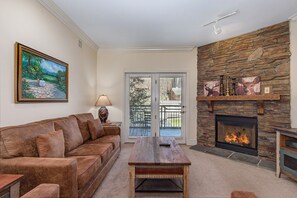 A living room with a stone fireplace, a sofa, a coffee table, and a painting on the wall. - Baskins Creek 213 🌄 Peaceful Balcony Views | Pool, Hot Tub & Fireplace 🏡 (Gatlinburg)