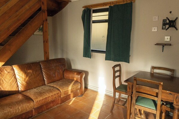 A room with a brown leather sofa, a wooden dining table with chairs, a window with curtains, and a wooden staircase.