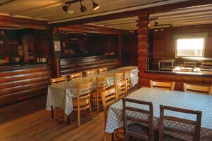 Un interior rústico de madera con mesas y sillas, una zona de cocina y un ventanal que proporciona luz natural.