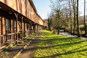 A wooden building with a covered walkway and a grassy area with a stream. - Cabin No. 14 - Salt and Fern Cabins (Port Talbot)