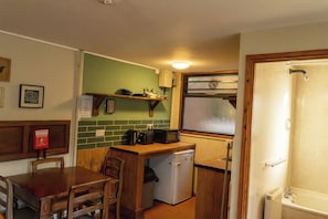 A compact kitchen with a refrigerator, microwave, and a small sink. There is a wooden table and chairs, and a window with a view. - Cabin No. 14 - Salt and Fern Cabins (Port Talbot)