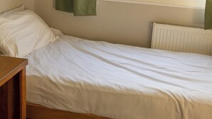 Egy egyágyas ágy, fa kerettel, fehér matraccal, és egy zöld törölköző lóg a falon.