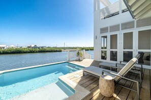 Ein Poolbereich mit Blick auf das Wasser, eine Holzterrasse, Liegestühle und ein Gebäude mit großen Fenstern.