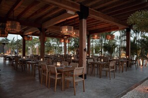An outdoor dining area with wooden tables and chairs, hanging lights, and a view of palm trees. - Abebi Oasis & Mistico Beach Club + Boat Shuttle (San Pedro)