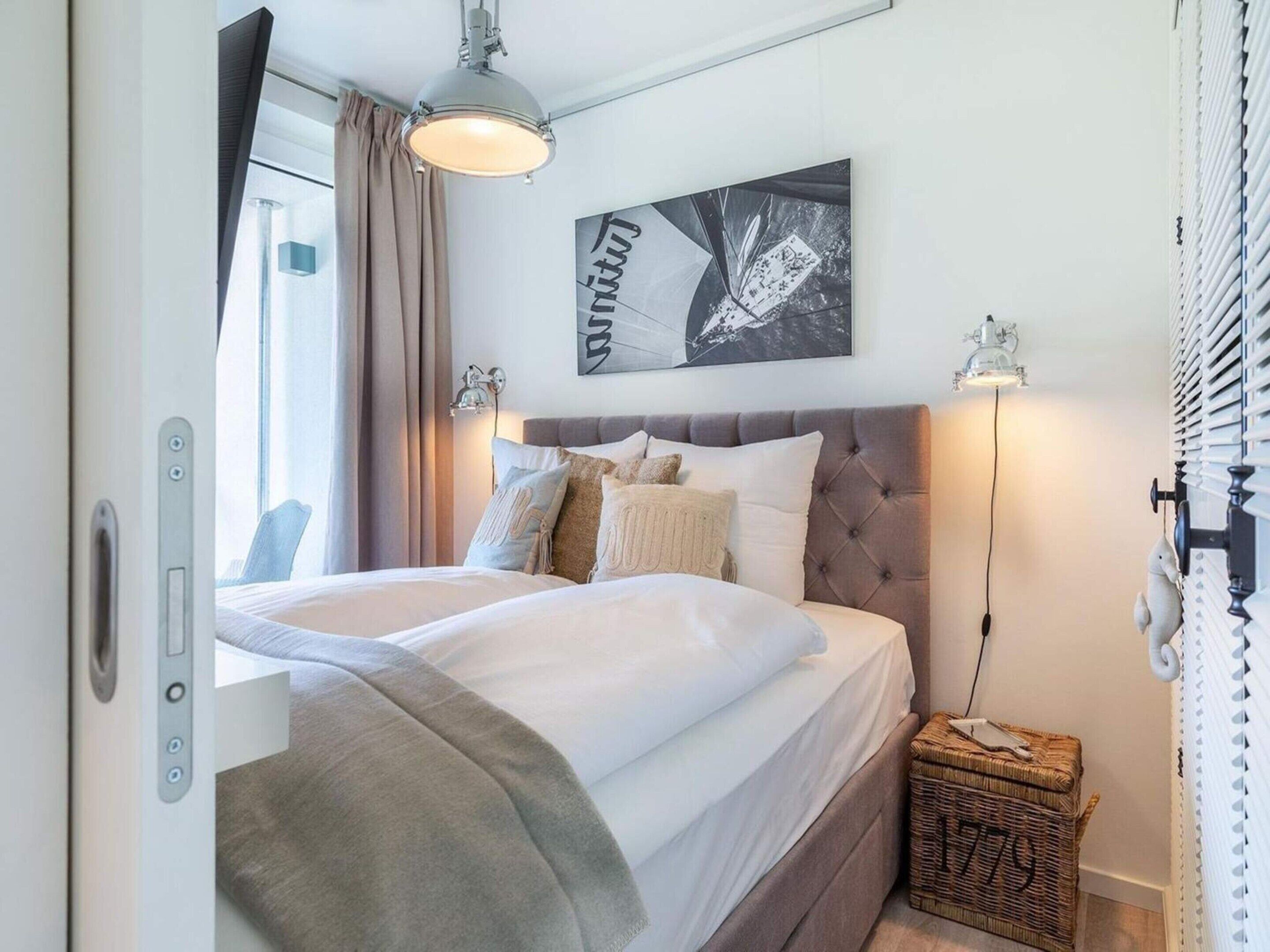 A bedroom with a bed, a headboard, a wall-mounted artwork, a bedside lamp, a window with curtains, and a wicker basket.