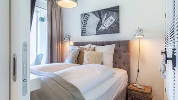A bedroom with a bed, a headboard, a wall-mounted artwork, a bedside lamp, a window with curtains, and a wicker basket.