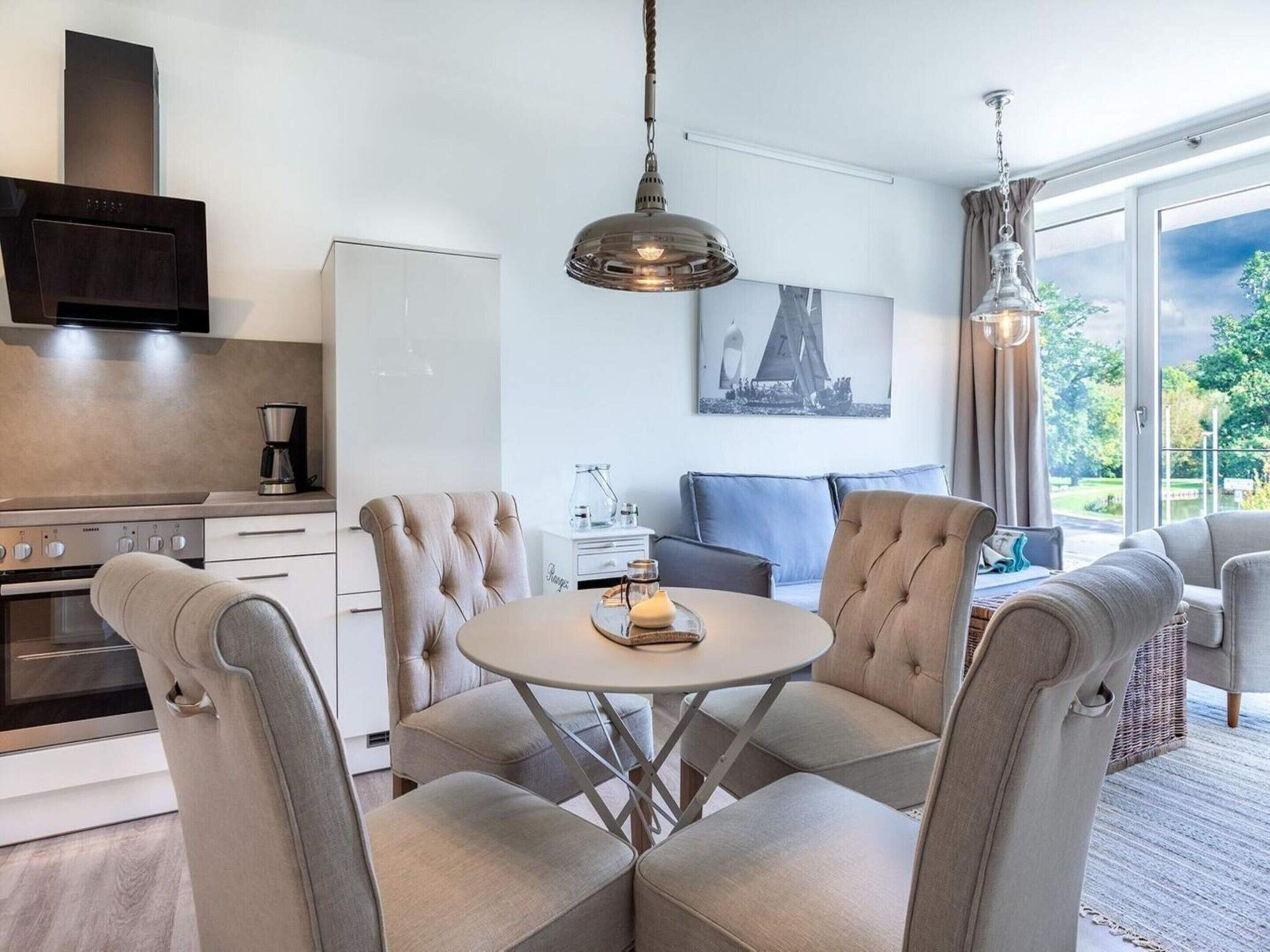 A modern kitchen with a built-in oven, a dining area with a round table and four chairs, a sofa, and a large window with a view of greenery.