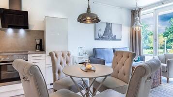 A modern kitchen with a built-in oven, a dining area with a round table and four chairs, a sofa, and a large window with a view of greenery.