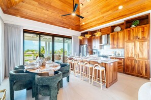 A modern kitchen with wooden cabinets, a central island, and a dining area with round tables and chairs. - BelaDia at Mahogany Bay - Luxurious 10 Bedroom Compound (San Pedro)