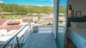 A balcony with a view of a town and hills, a bed with a view, and a glass door. - Travessa Apartments 3 Bedrooms By PontaNegraAzores (Praia da Vitória)
