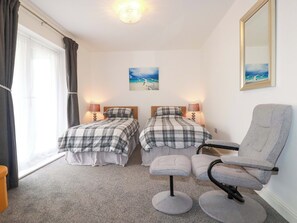 Una habitación con dos camas, un sillón gris, un taburete pequeño, un cuadro y un espejo.