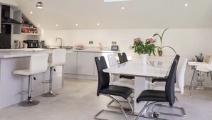 Una cocina moderna con tragaluces, taburetes de bar blancos, una mesa de comedor con sillas y una mesita auxiliar con un jarrón de flores.