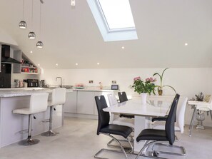 Una cocina moderna con tragaluces, taburetes de bar blancos, una mesa de comedor con sillas y una mesita auxiliar con un jarrón de flores.