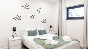 A modern bedroom with a white bed, green throw, and towels, a nightstand with a lamp, and origami wall art. - Travessa Apartments 1 Bedroom By PontaNegraAzores (Praia da Vitória)