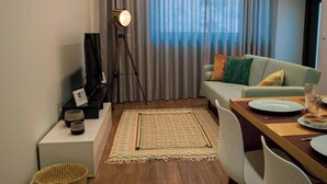 Ett vardagsrum med en soffa, ett soffbord, ett matbord med stolar, en lampa och en utsikt genom gardinerna.