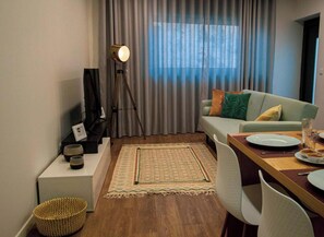 Ein Wohnzimmer mit einer Couch, einem Couchtisch, einem Esstisch mit Stühlen, einer Lampe und Blick durch Vorhänge.