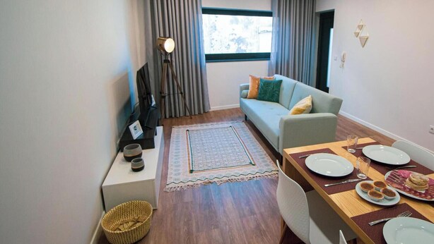 A modern living room with a sofa, dining table set for two, and a large window with curtains. - Travessa Apartments 1 Bedroom By PontaNegraAzores (Praia da Vitória)