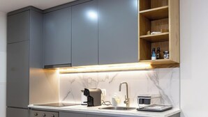 A modern kitchen with grey cabinets, a marble countertop, a built-in oven, a coffee machine, a sink, and a wooden shelf with bottles. - Travessa Apartments 1 Bedroom By PontaNegraAzores (Praia da Vitória)