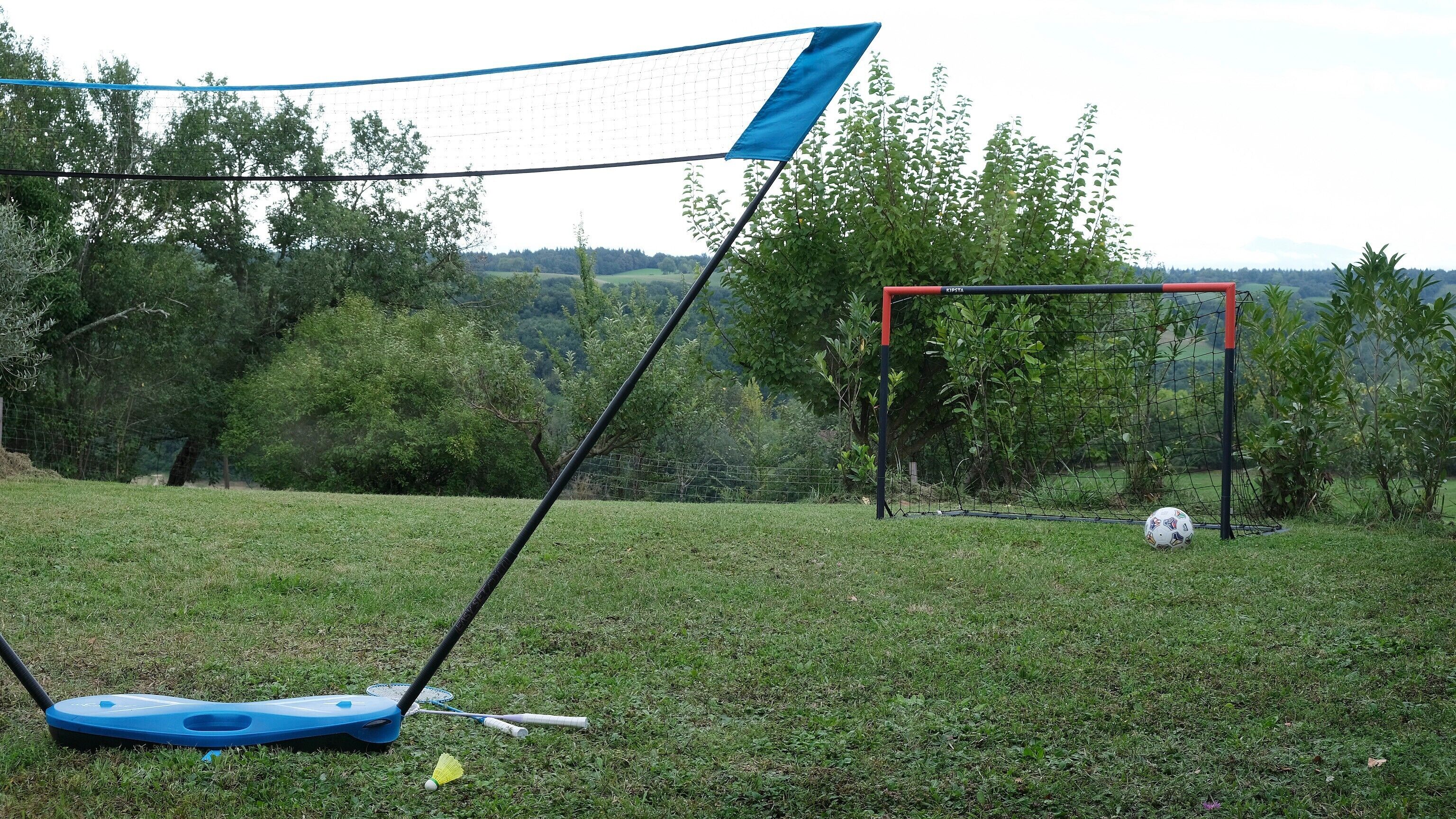 Een badmintonnet, een voetbalkooi, en een gazon met bomen op de achtergrond.
