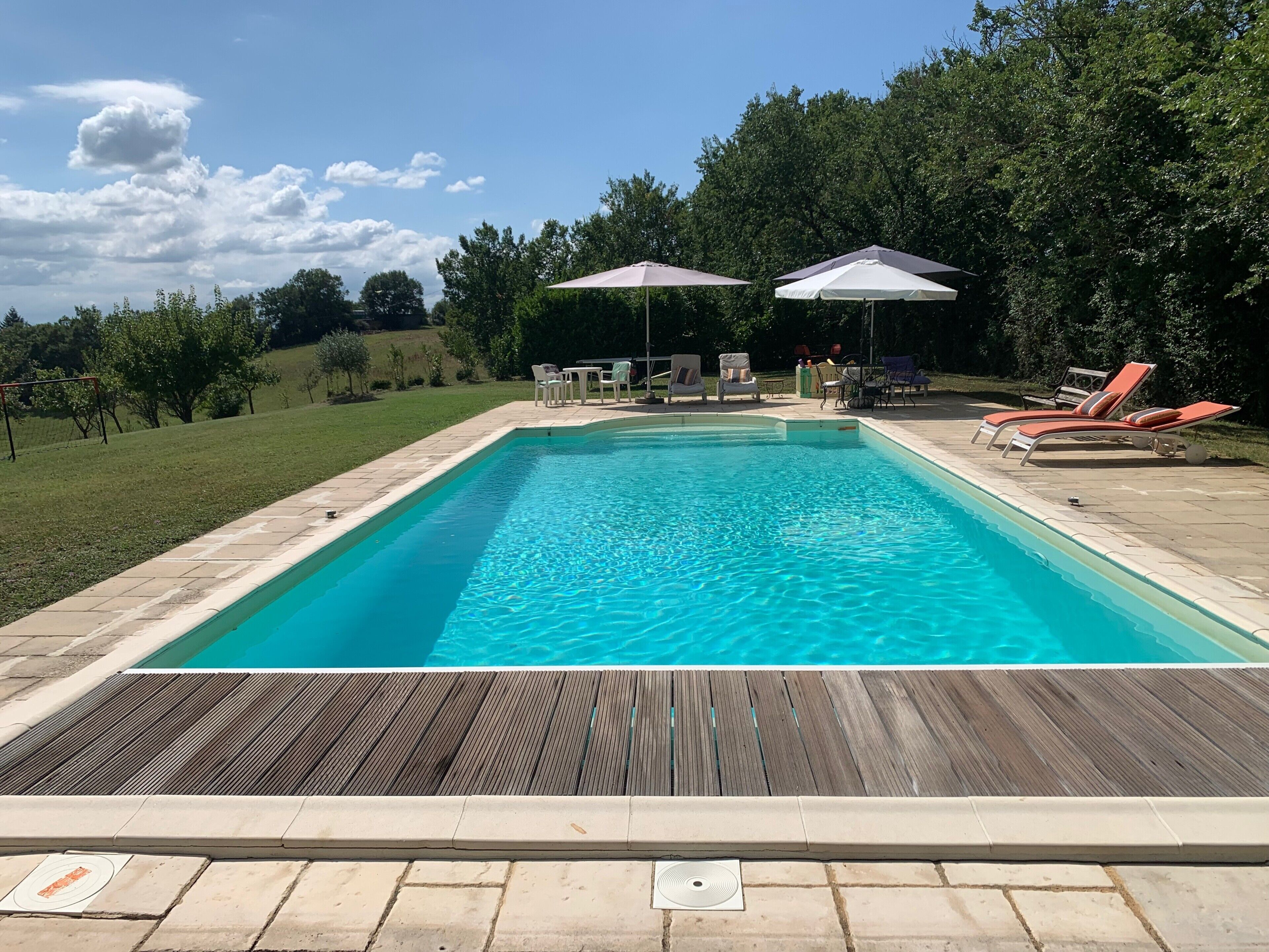 Ein rechteckiger Swimmingpool mit Holzterrasse im Vordergrund, umgeben von Liegestühlen und Sonnenschirmen.