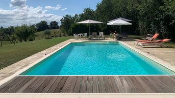 Ein rechteckiger Swimmingpool mit Holzterrasse im Vordergrund, umgeben von Liegestühlen und Sonnenschirmen.