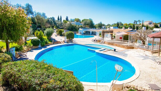 Una piscina amplia y cristalina con una terraza soleada y sillones de descanso, rodeada de vegetación y edificios residenciales.