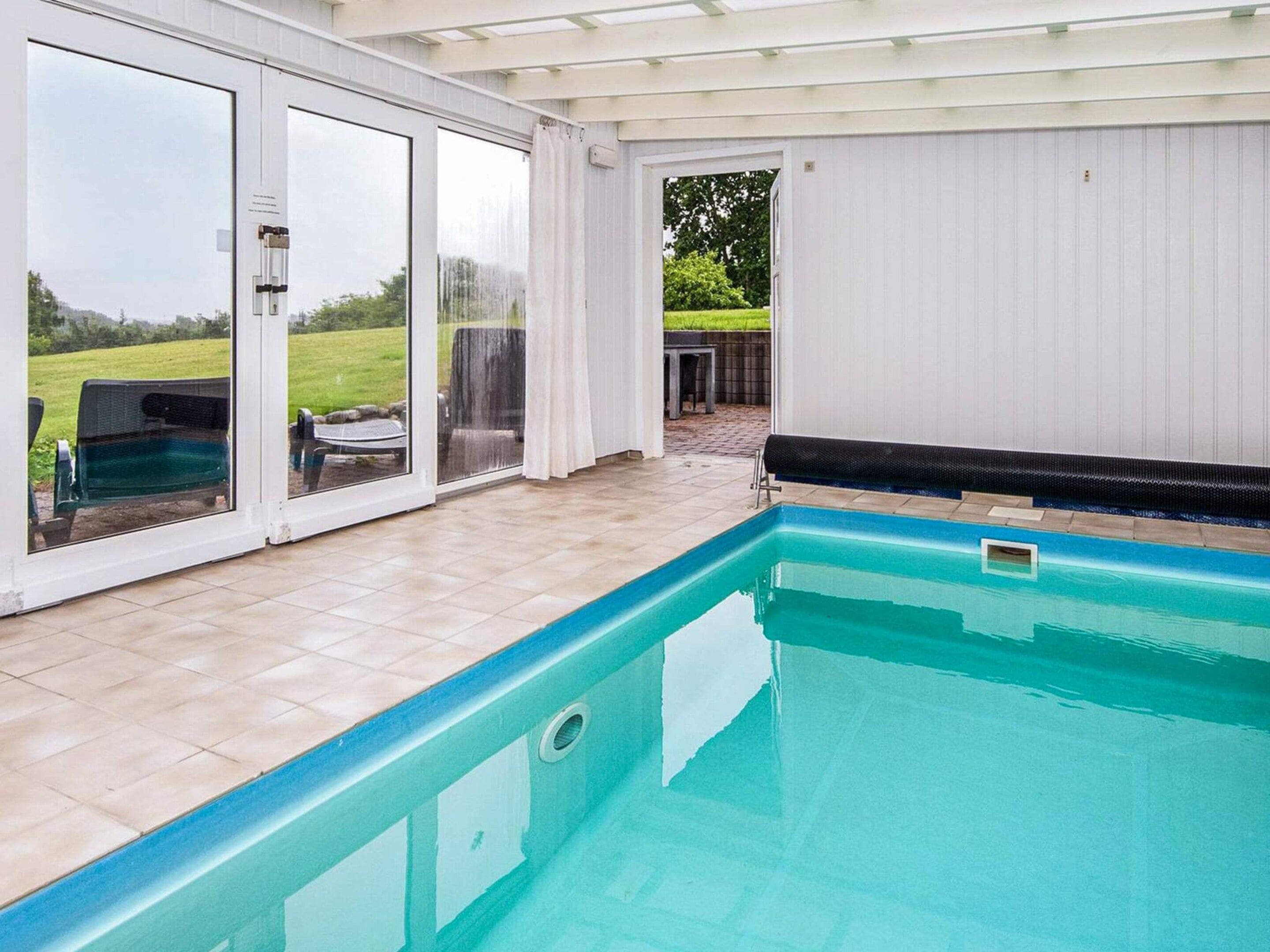 A swimming pool with a tiled deck area, a covered patio with a view of the outdoors, and a glass door leading to an outdoor seating area.