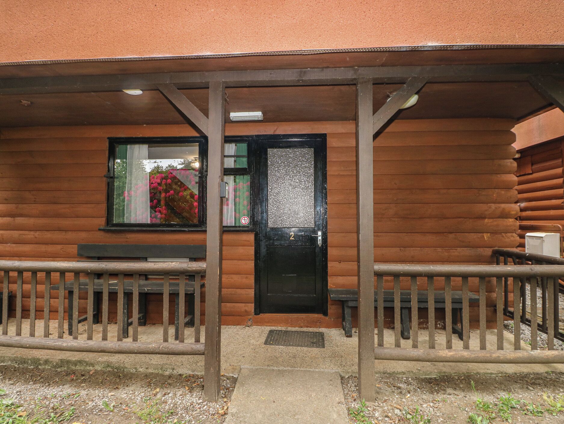 Eine Blockhütte mit einer schwarzen Tür, einem Fenster mit Blick auf rosa Blumen und einer Bank davor.