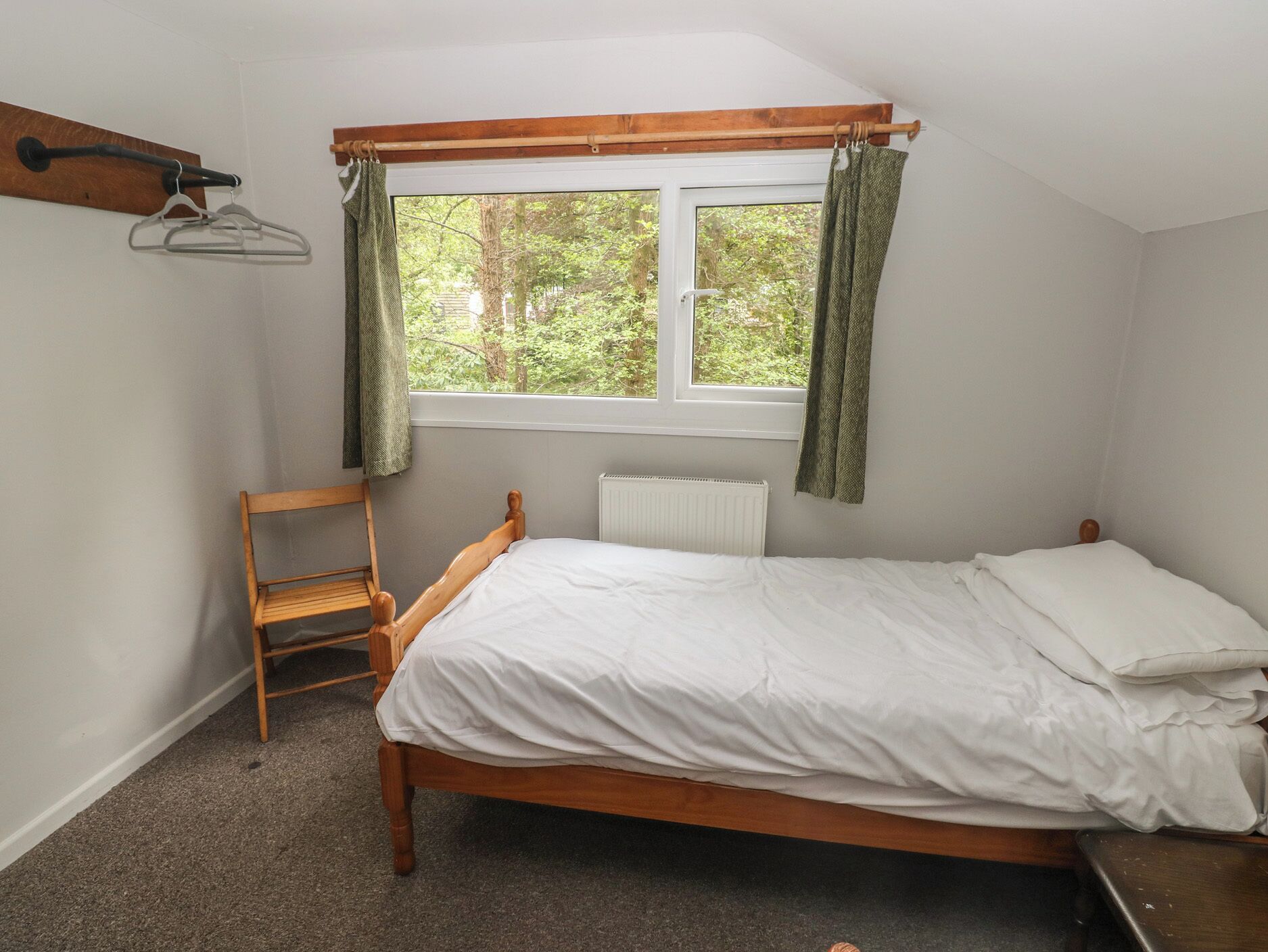 Ein Einzelbett mit Holzrahmen, ein Heizkörper unter dem Fenster, ein Holzstuhl und ein Kleiderständer mit Kleiderbügeln.