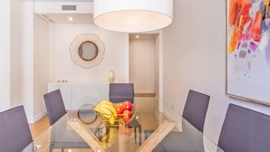 Un comedor moderno con una mesa de vidrio, sillas de estructura metálica, una lámpara colgante blanca y un cuadro abstracto colorido.