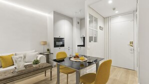 A modern living room with a sofa, a dining table with chairs, a kitchen area, and a white door. - Cozy Studio by Colón & Salamanca District (Madrid)