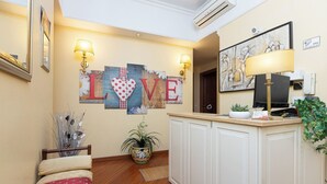 Hotelkamer met houten vloer, een bed met beddengoed met patroon, een receptiebalie met lamp, een muur met de tekst 'LOVE' en een schilderij, en een bloempot met plant.