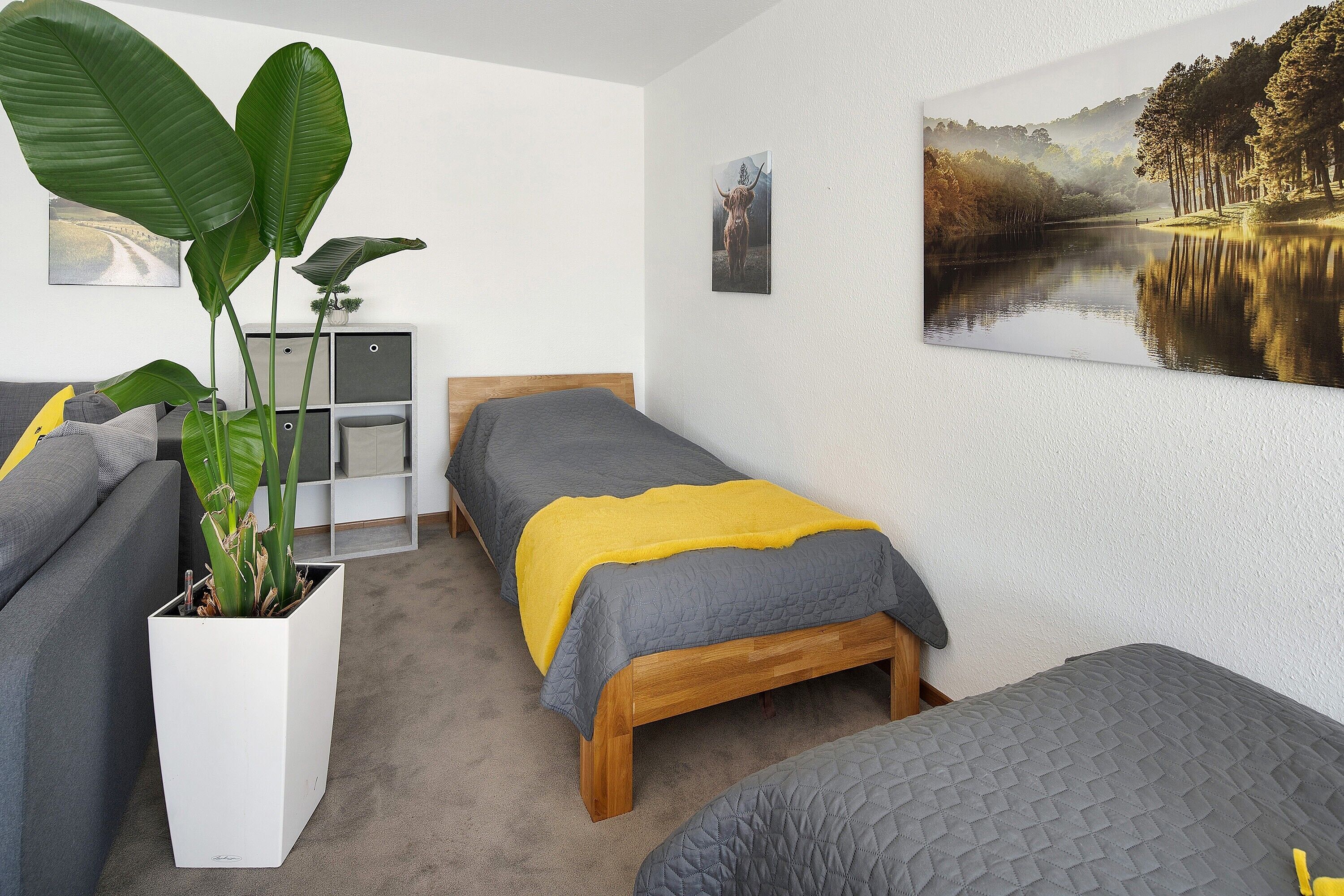 Un dormitorio con dos camas, una planta grande en maceta y un cuadro en la pared.