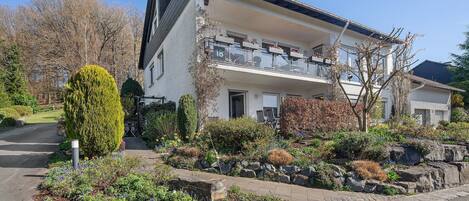 A two-story residential building with a balcony, a stone retaining wall, and a landscaped garden.