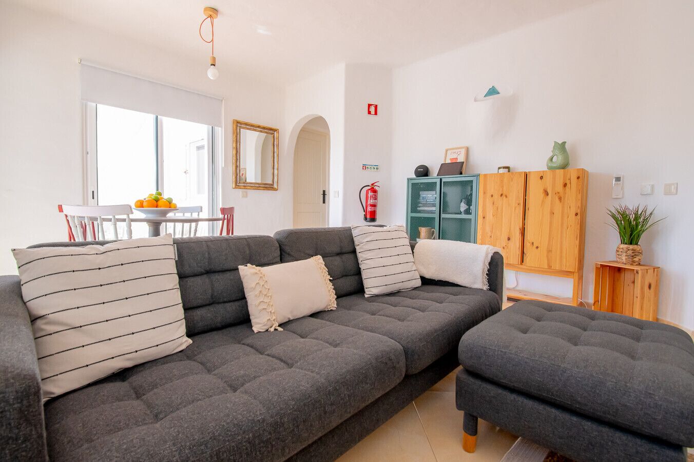 Un salon moderne avec un canapé d’angle gris, un petit pouf, un meuble en bois et un coin repas comprenant une table et des chaises.