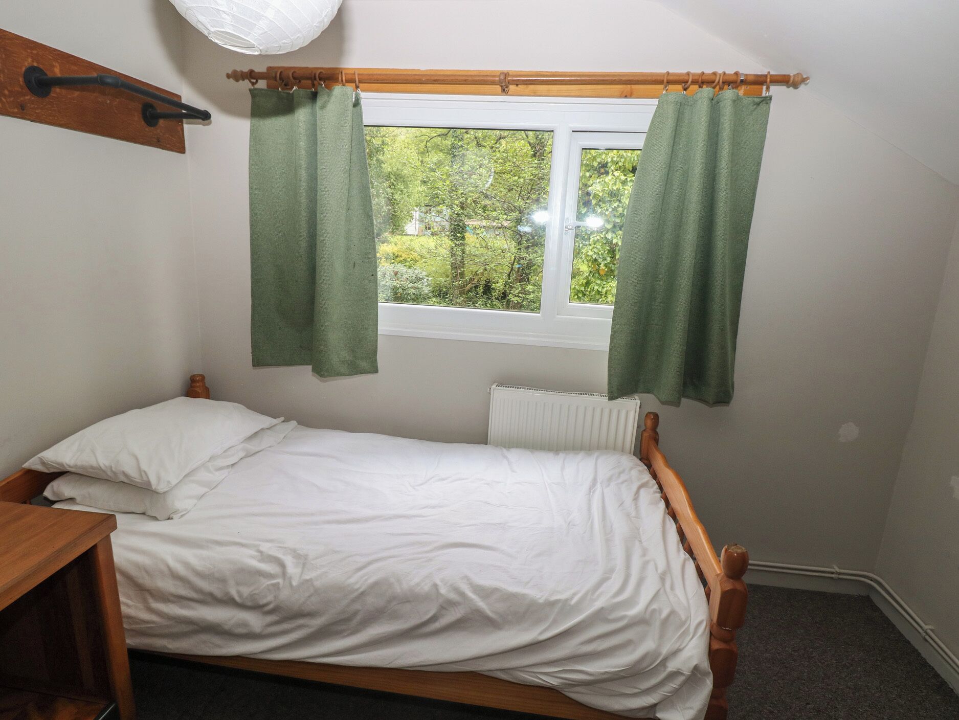 En säng med plats för en person, med vita sängkläder, ett träfärgat sänggavel, ett fönster med gröna gardiner, ett element under fönstret och en trähylla ovanför sängen.