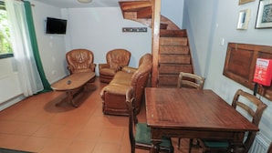 Una habitación con una escalera de madera, una mesa de comedor con sillas, dos sillones de cuero, una mesita pequeña y un televisor montado en la pared.