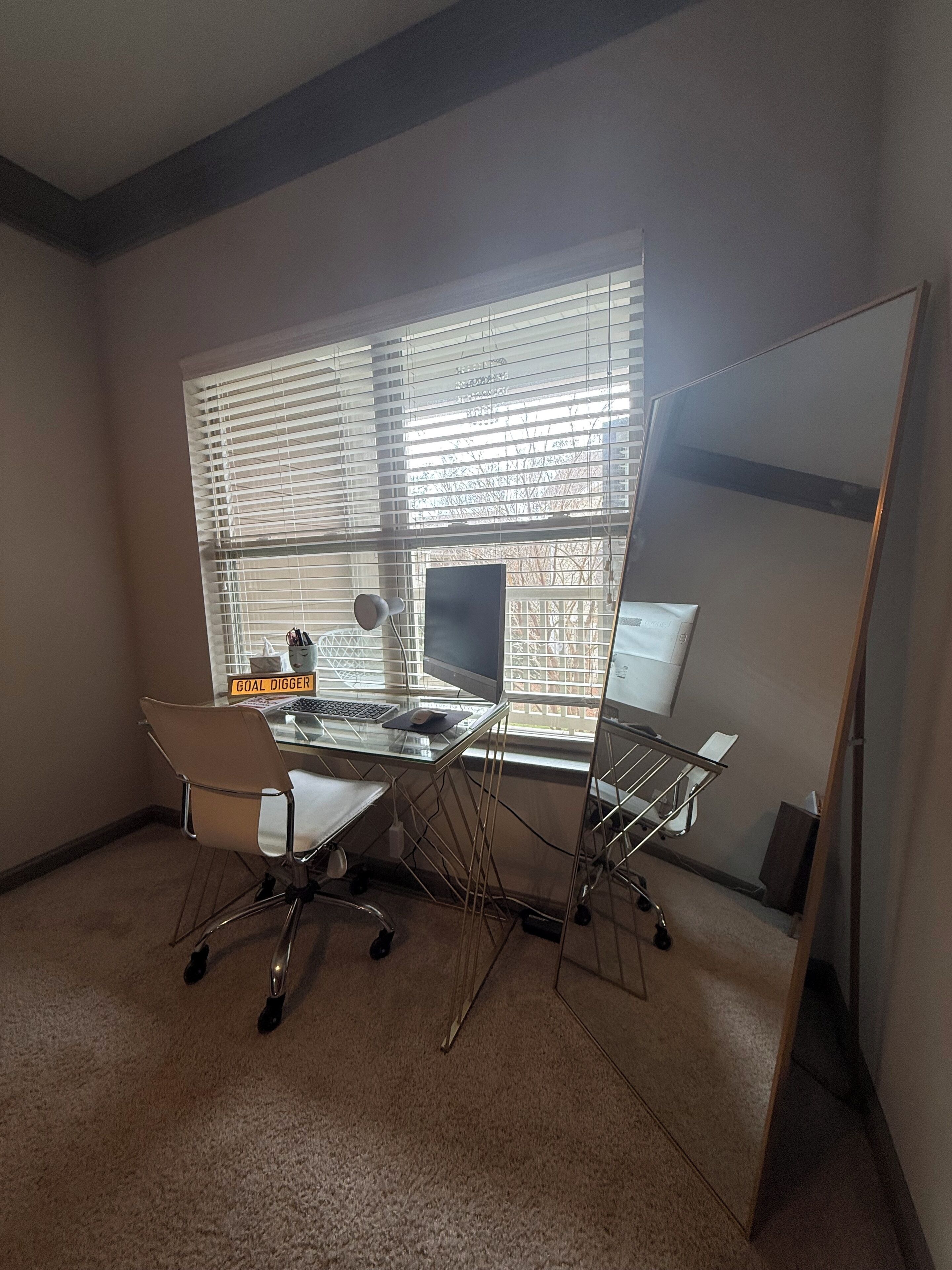 Ein Zimmer mit Schreibtisch, Computer, Stuhl und einem großen Spiegel.