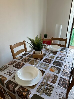 Ein Esstisch mit einer Tischdecke mit Kaffeemotiv, einer Topfpflanze und einem Geschirrset aus weißen Tellern und Besteck.