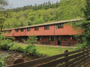 A two-story building with a red exterior, surrounded by lush greenery and a small stream. - Cabin No. 13 - Salt and Fern Cabins (Port Talbot)