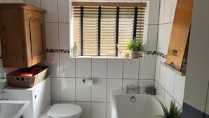Une salle de bain comprenant des toilettes, un lavabo, une baignoire, un meuble en bois, une fenêtre avec des stores et une plante en pot.