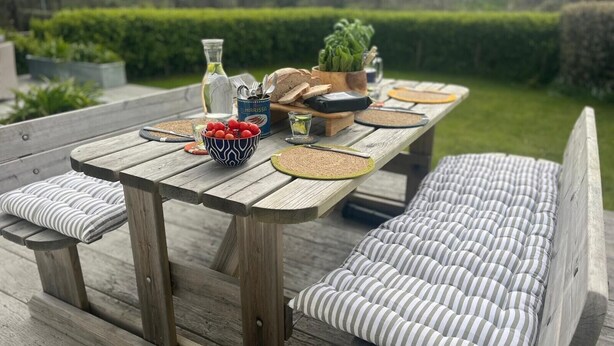 Une terrasse en bois avec une table dressée pour un repas, comprenant un saladier de fruits, une bouteille et une plante.