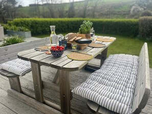 A wooden deck with a table set for a meal, including a bowl of fruit, a bottle, and a plant. - Fabulous three bed apartment with large exclusive contained garden and patio (Porthcurno)