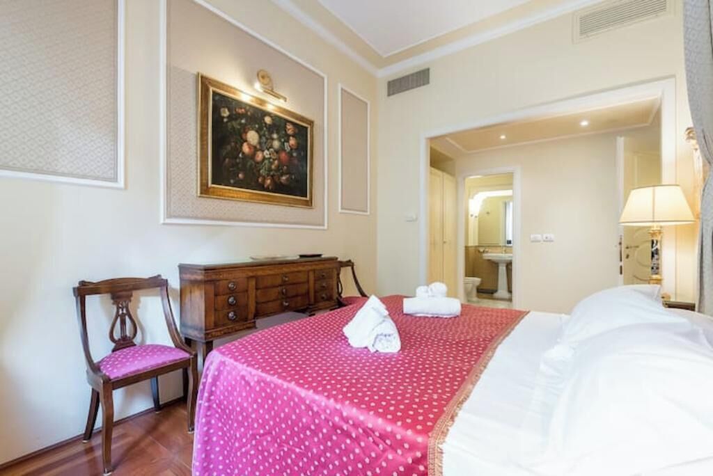 A hotel room with a bed, a chair, a dresser, a painting, and a bathroom visible through an open door.