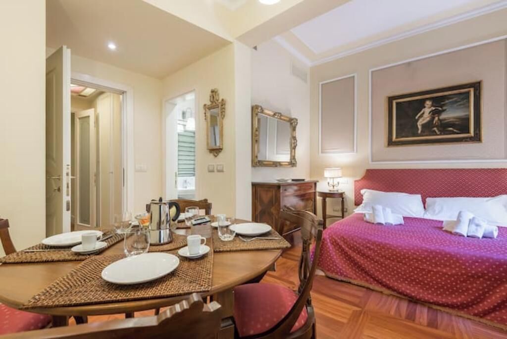 Een hotelkamer met een eettafel gedekt voor twee personen, een bed met een rode plaid, een schilderij aan de muur en een spiegel boven het bed.