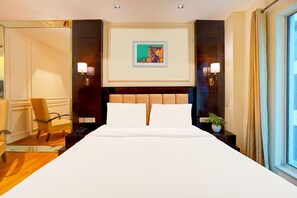 A hotel room with a large bed, a chair, a nightstand with a lamp, a framed picture on the wall, and a window with curtains.