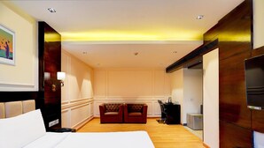 A hotel room with a large bed, a wooden headboard, a seating area with two leather sofas, a TV, and a minibar. - Deluxeimperial Villa Delhi (New Delhi)