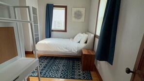 Um quarto pequeno e bem organizado, com uma cama beliche, uma cama de solteiro, um tapete estampado e uma janela com cortinas.