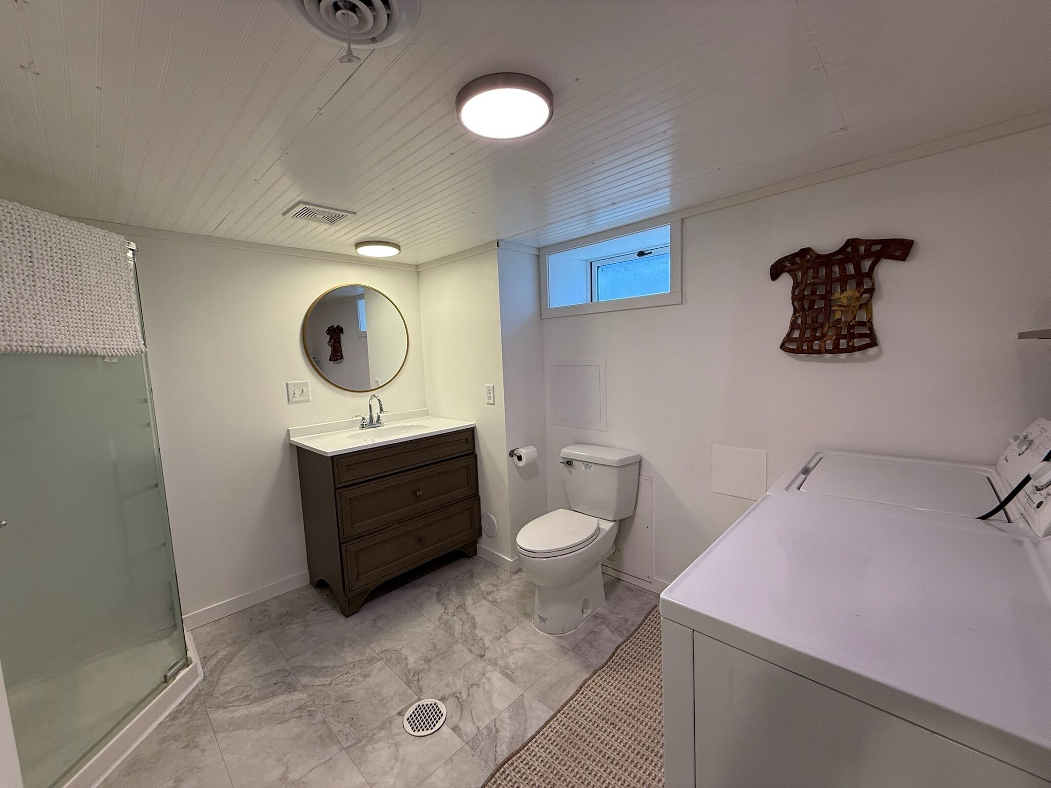 A bathroom with a glass shower enclosure, a white sink with a round mirror, a dark wooden vanity, a toilet, and a white bathtub.
